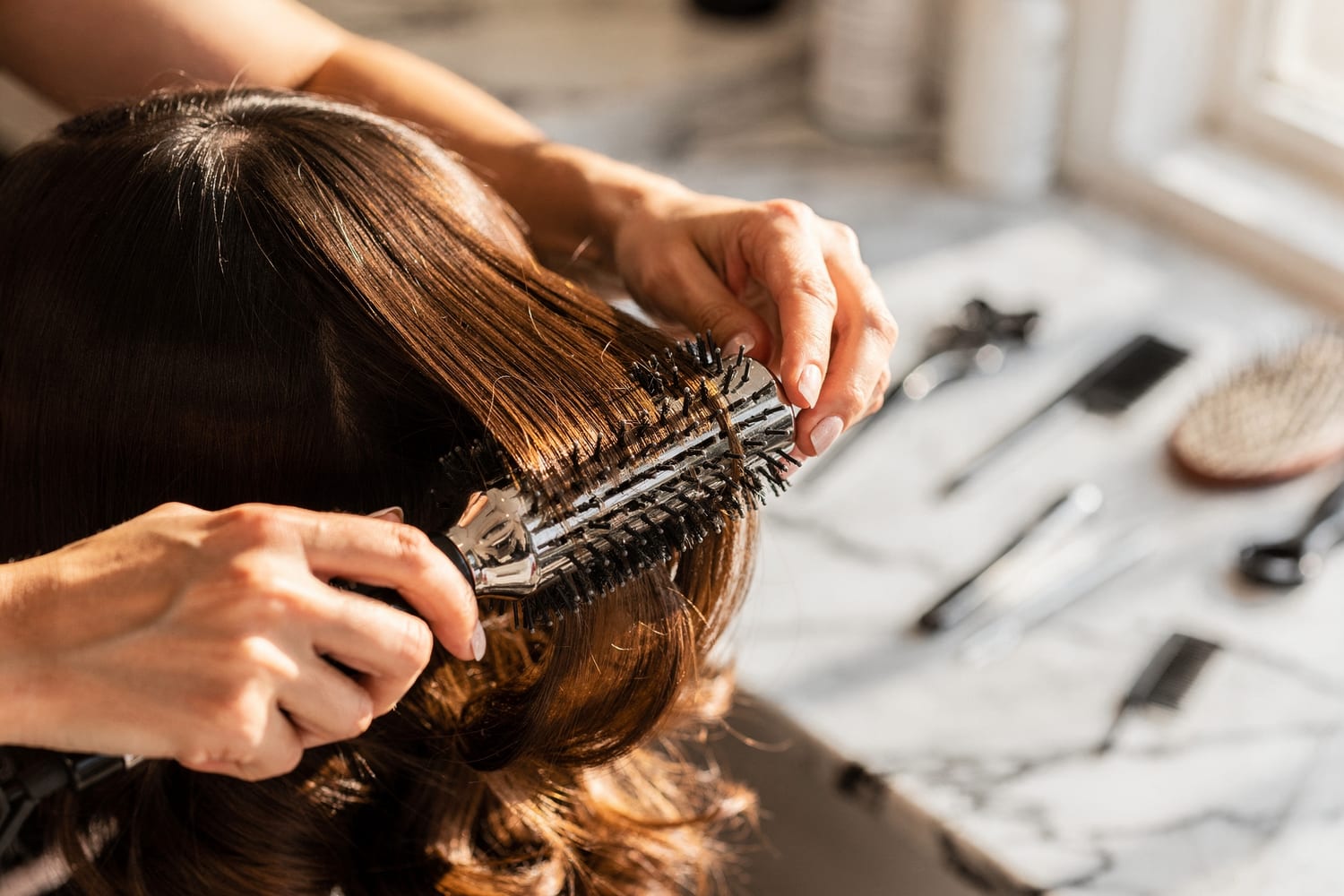 A stylist finishing glossy, styled hair with a round brush on a marble station
